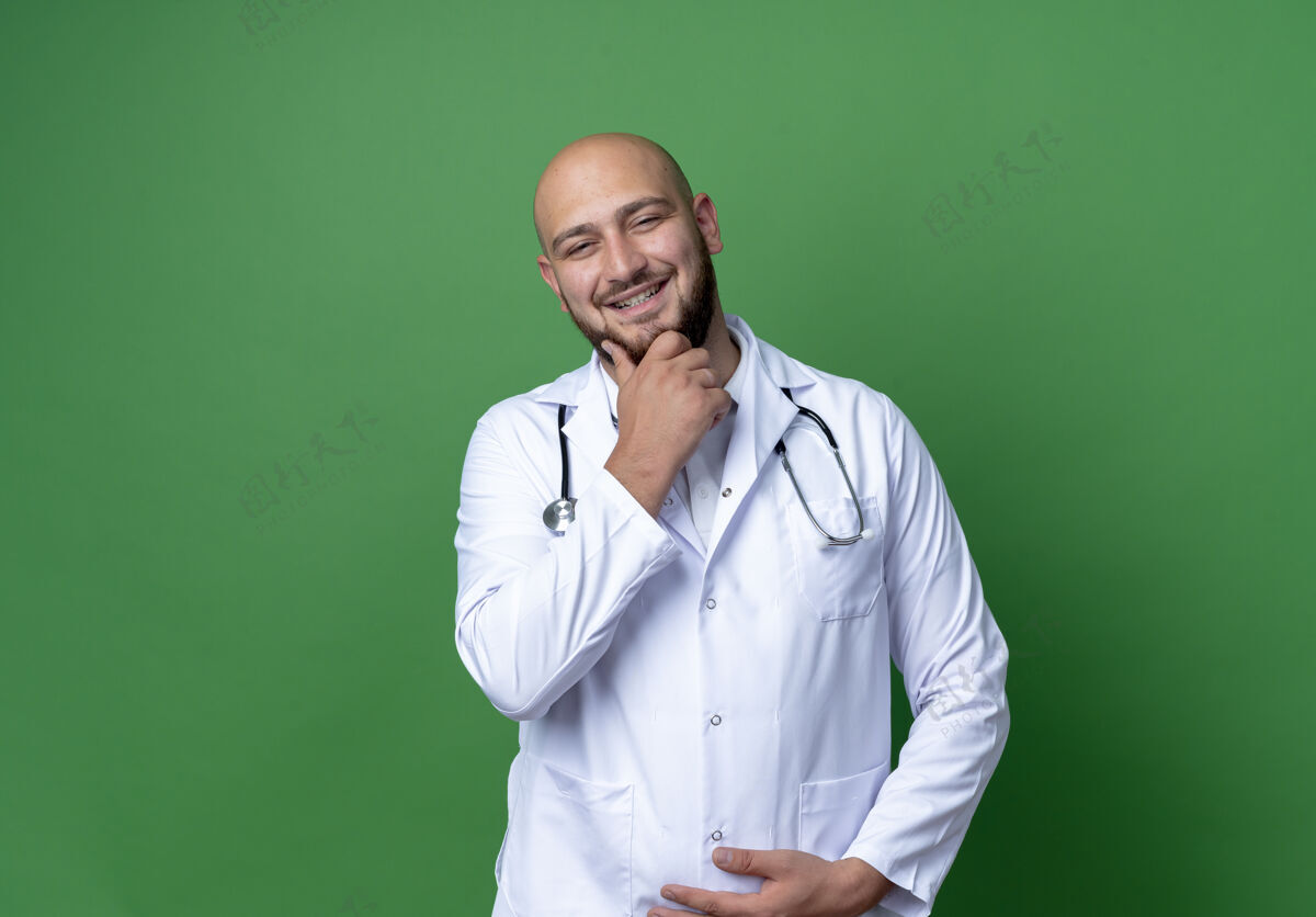 微笑微笑的年轻男医生穿着医用长袍和听诊器 手放在绿色背景上孤立的下巴上手戴医生