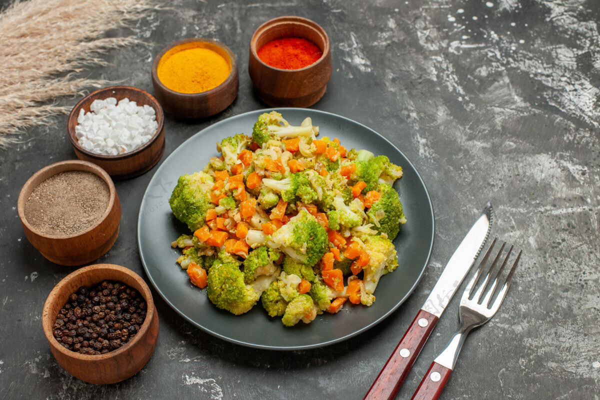 午餐健康餐的侧视图 黑色盘子上有花椰菜和胡萝卜 灰色桌子上有香料晚餐锅壁板