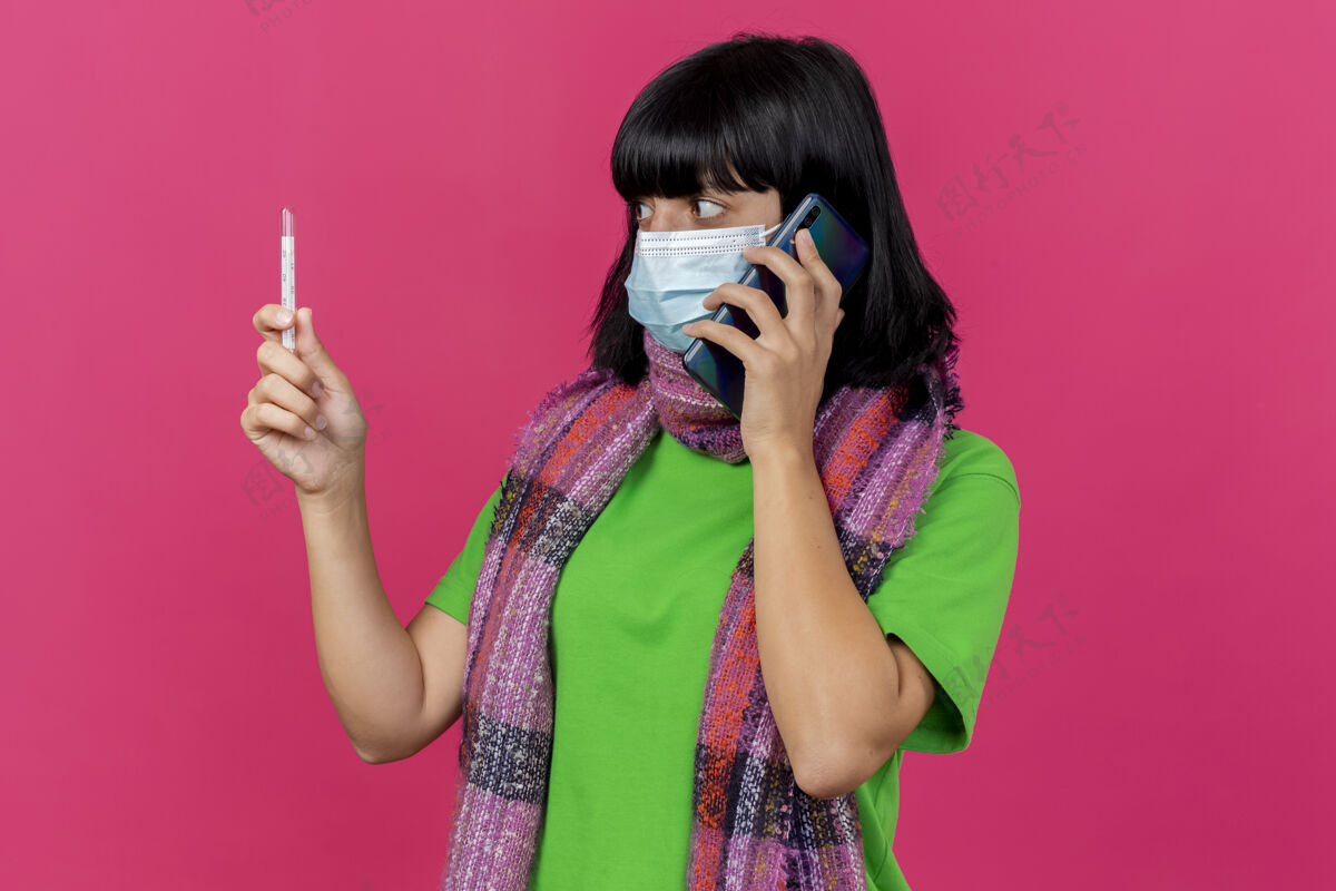 体温计印象深刻的年轻生病的女人戴着口罩和围巾拿着和看温度计讲电话隔离在粉红色的墙壁上复制空间年轻人公民站着