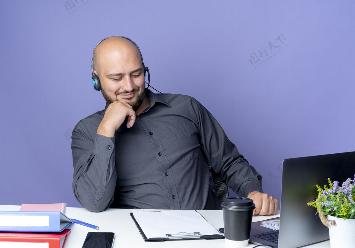 穿高兴的年轻秃顶呼叫中心男子戴着耳机坐在办公桌旁 手放在下巴上 手放在紫色背景上 看着剪贴板耳机电话中心