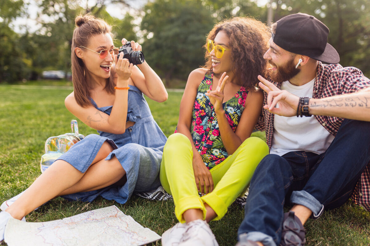 积极快乐的年轻人伴着谈笑风生的朋友坐在公园里 男女同乐 缤纷的夏日潮人时尚风格 带着相机旅行多种族休闲黑色
