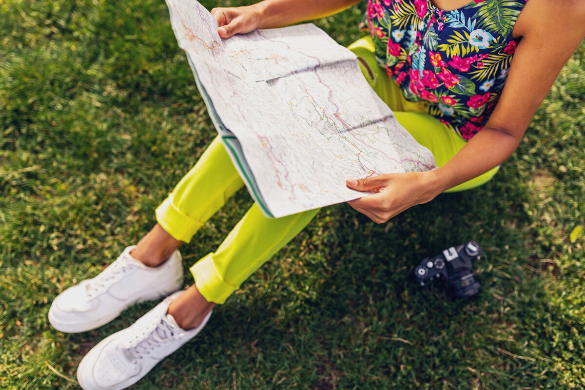 年轻女人手拿地图 旅行者拿着相机在公园里玩夏天的时尚风格 五颜六色的时髦装扮 坐在草地上 黄色的裤子明亮相机旅游