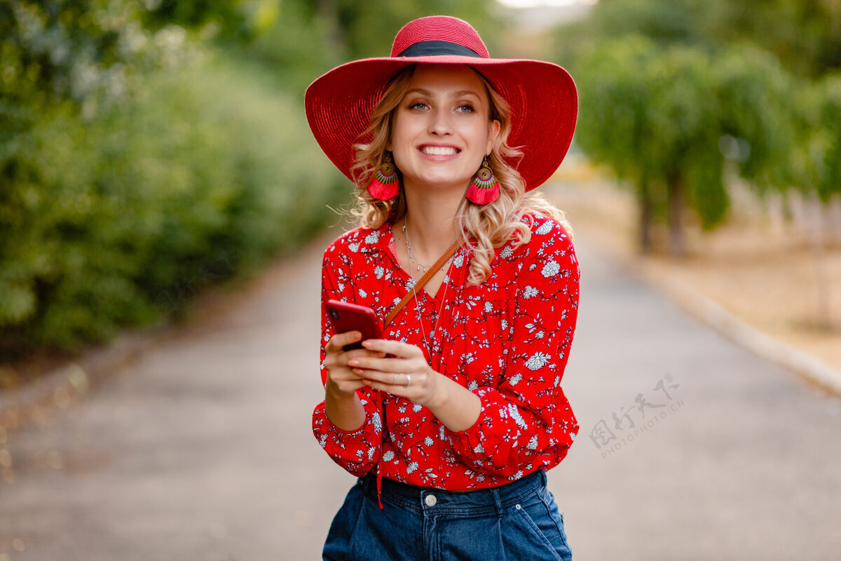 年轻迷人的时尚金发微笑的女人在稻草红帽子和衬衫夏季时尚套装使用手机配饰情感明亮