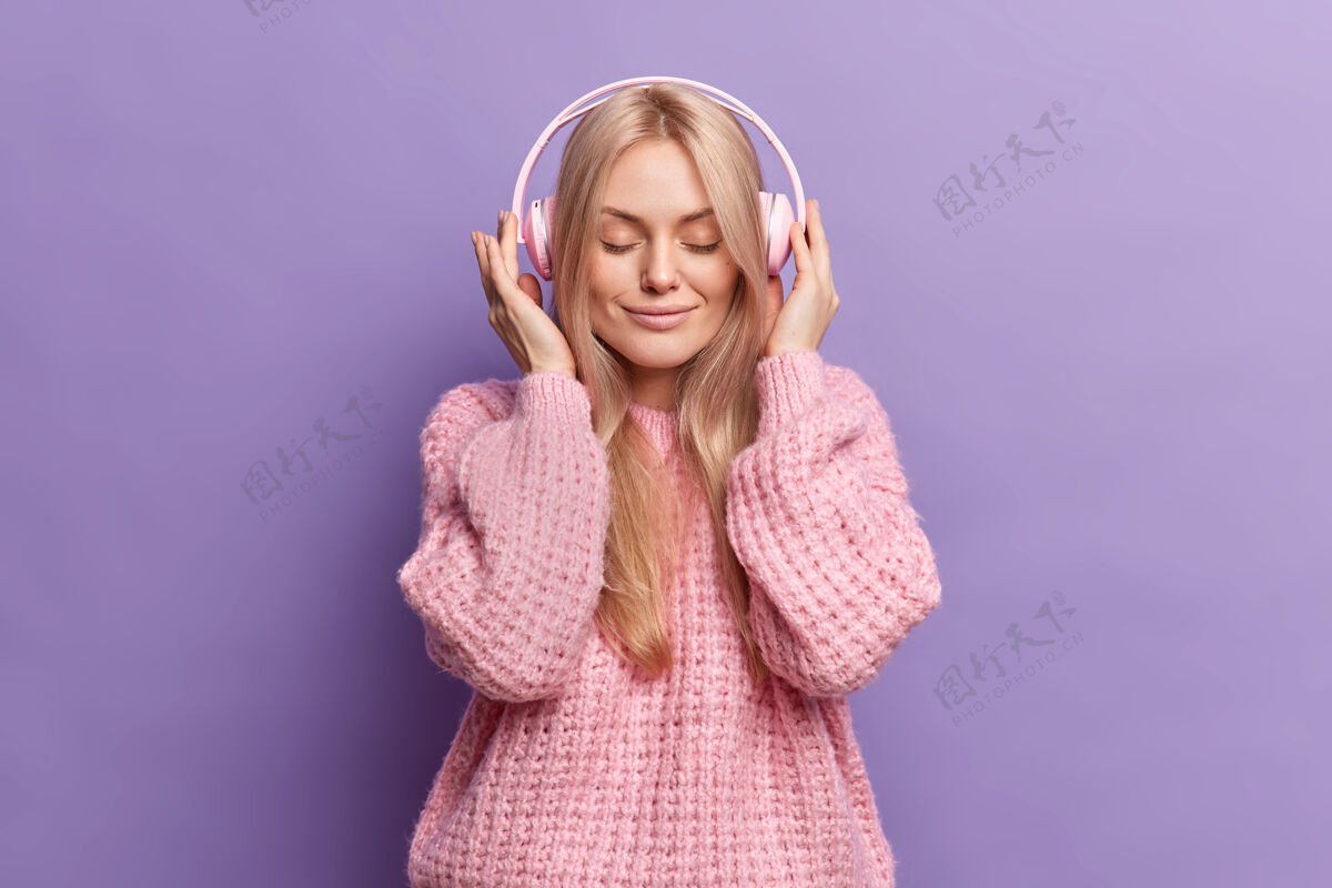 积极可爱冷静的金发女人手戴立体声耳机眼睛紧闭听着音乐穿着针织套头衫欣赏每一首曲子金发女性快乐