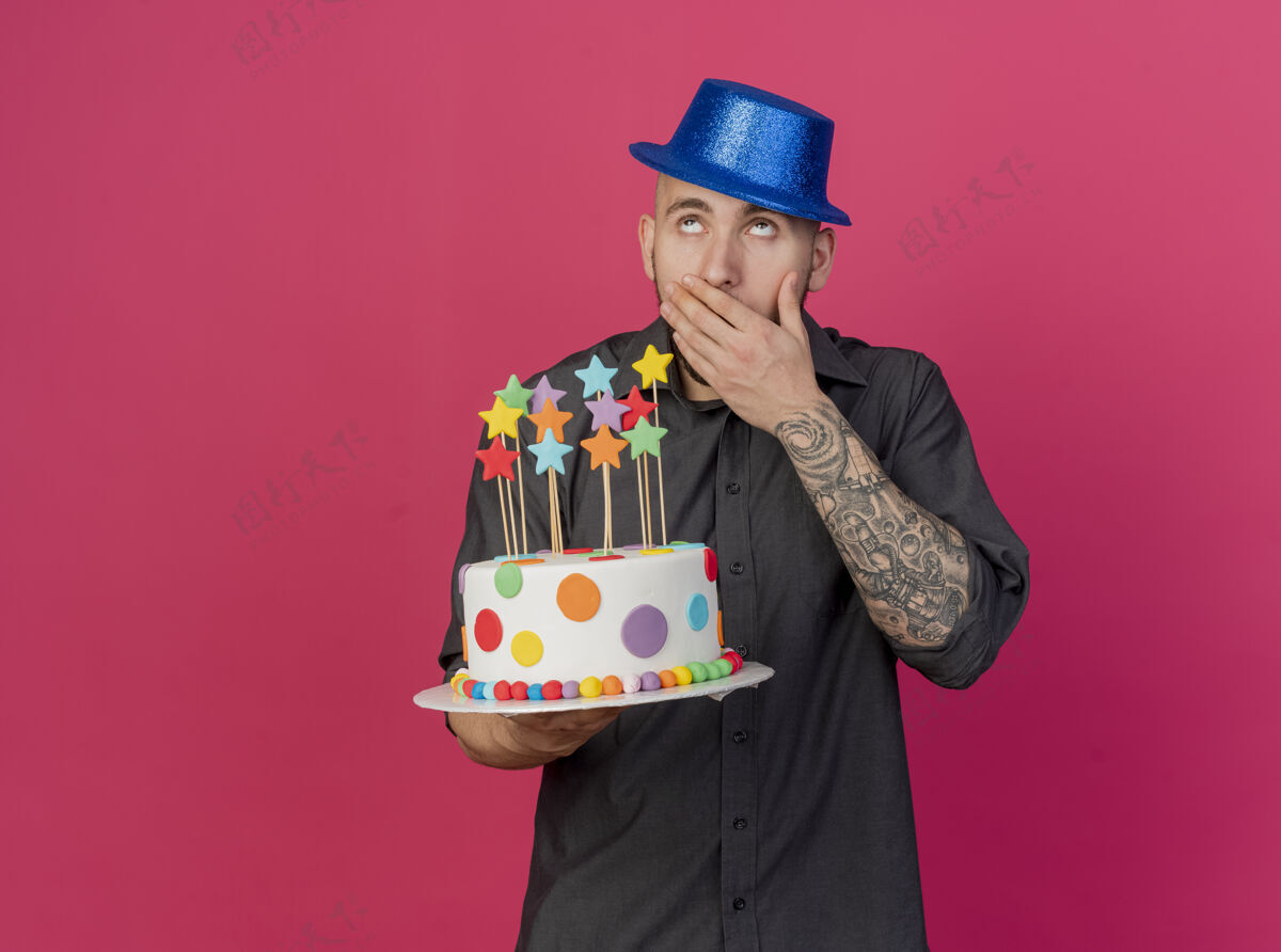 举行令人惊讶的年轻英俊的斯拉夫党人戴着党帽拿着生日蛋糕与明星仰望保持手放在嘴上红色背景与复制空间隔离复制派对星星