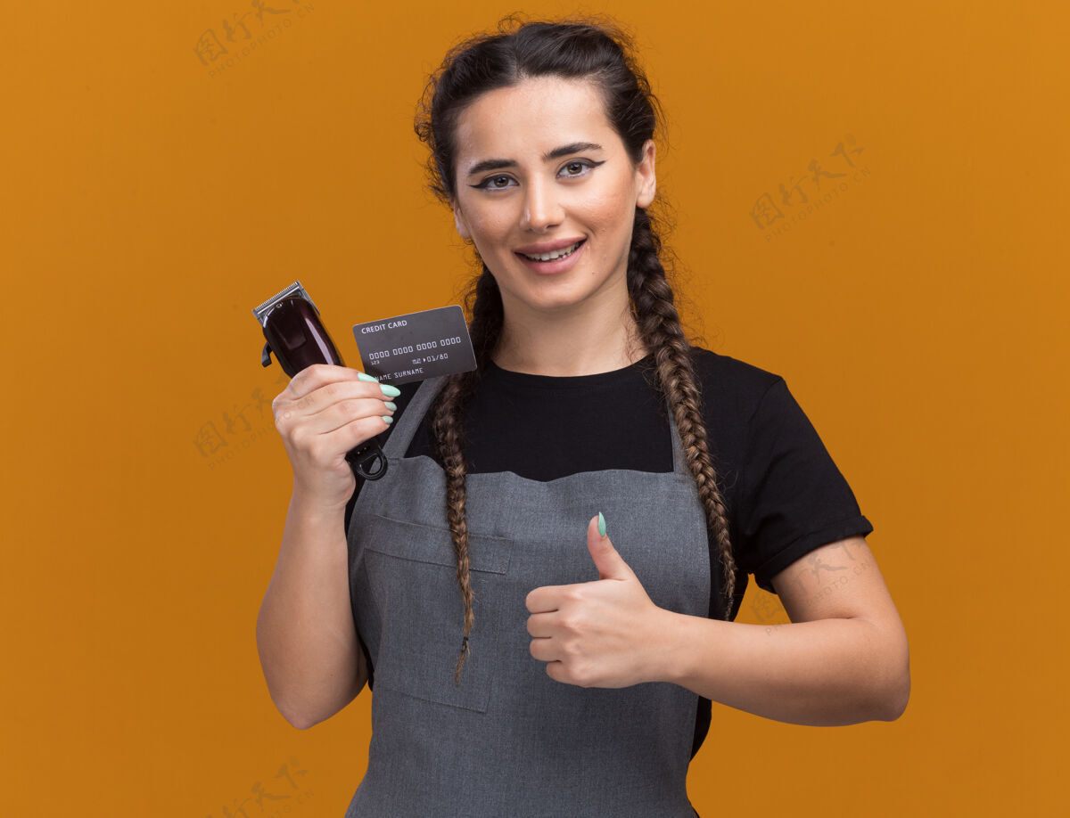 年轻微笑着的年轻女理发师穿着制服拿着信用卡和理发器在橙色的墙上孤立地竖起大拇指持有女信用卡