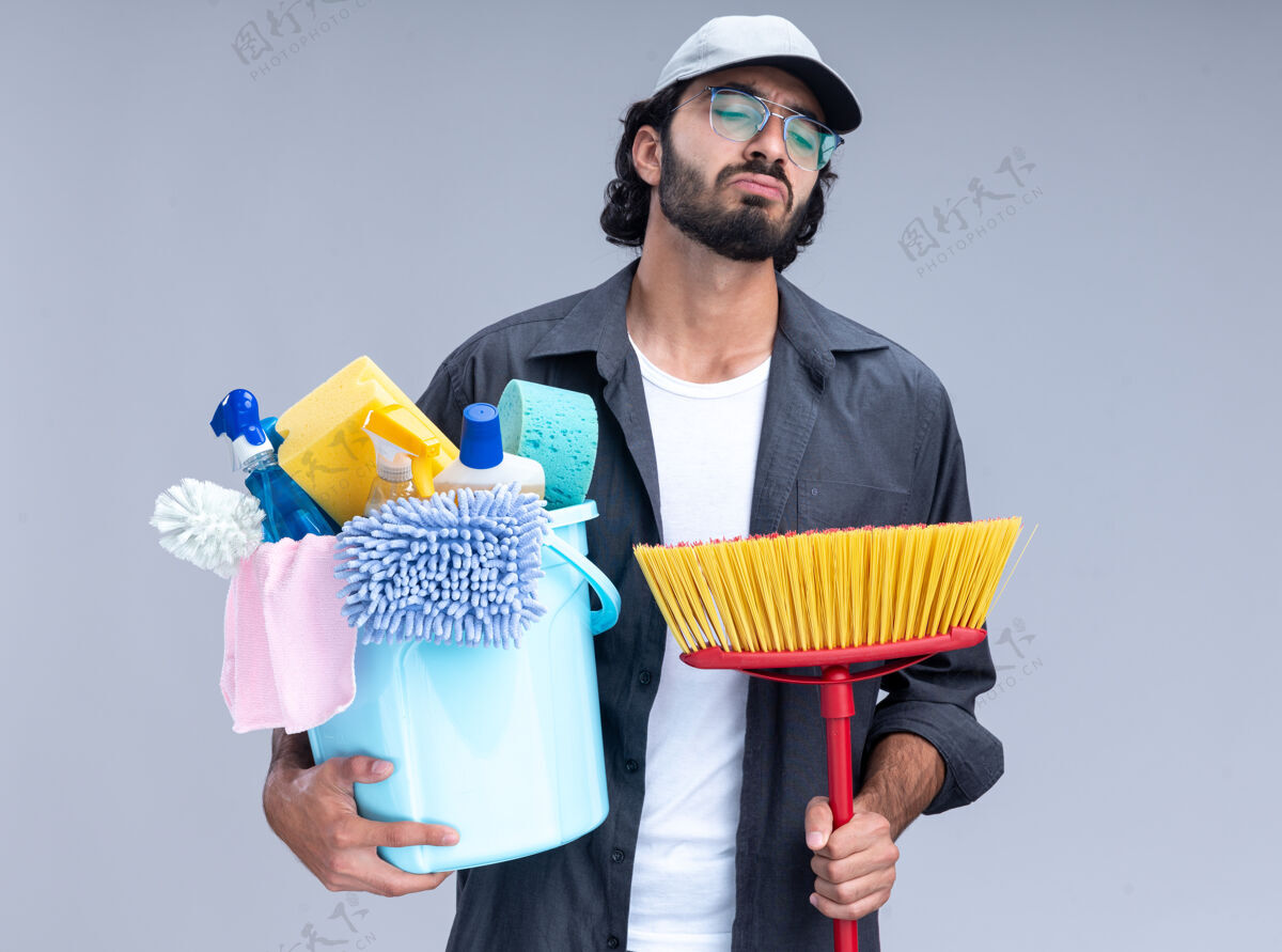 T恤悲伤的年轻帅气的清洁工穿着t恤 戴着帽子 手里拿着一桶清洁工具和拖把 隔离在白色的墙上清洁家伙水桶