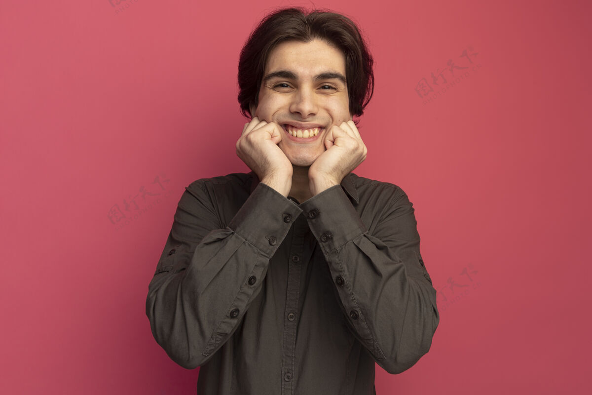 穿面带微笑的年轻帅哥 穿着黑色t恤 手放在粉色墙上孤立的脸颊上年轻T恤手