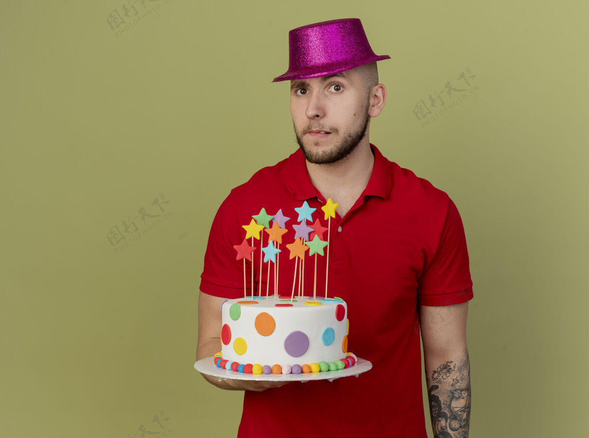 抱着可疑的年轻帅哥斯拉夫党家伙戴着党的帽子拿着生日蛋糕看着相机孤立在橄榄绿的背景与复制空间复制空间蛋糕