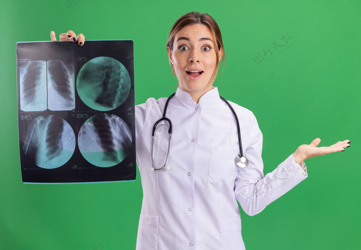 年轻年轻的女医生穿着医用长袍 手持听诊器 手拿着x光片 被隔离在绿墙上穿长袍传播
