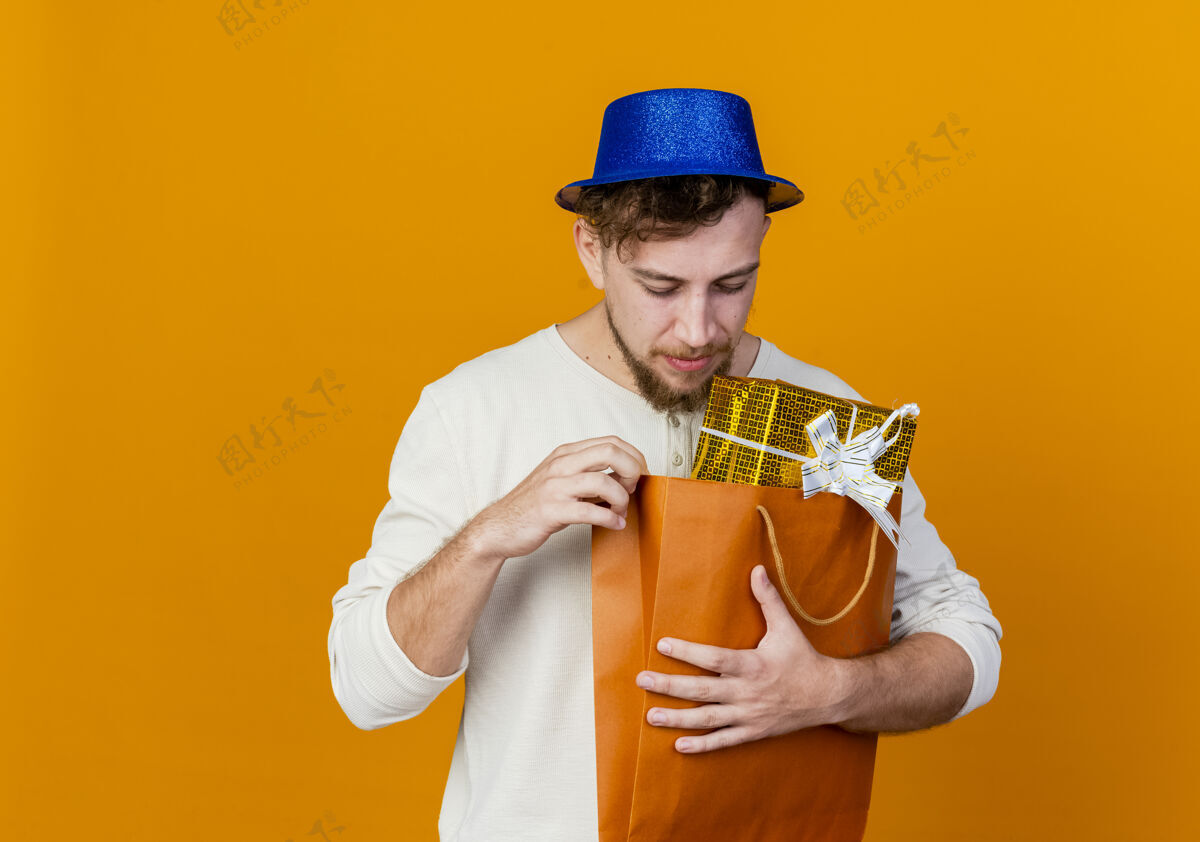 派对年轻英俊的斯拉夫党人戴着党的帽子举行 并期待与礼品盒内的橙色背景与复制空间隔离纸袋礼物盒子内
