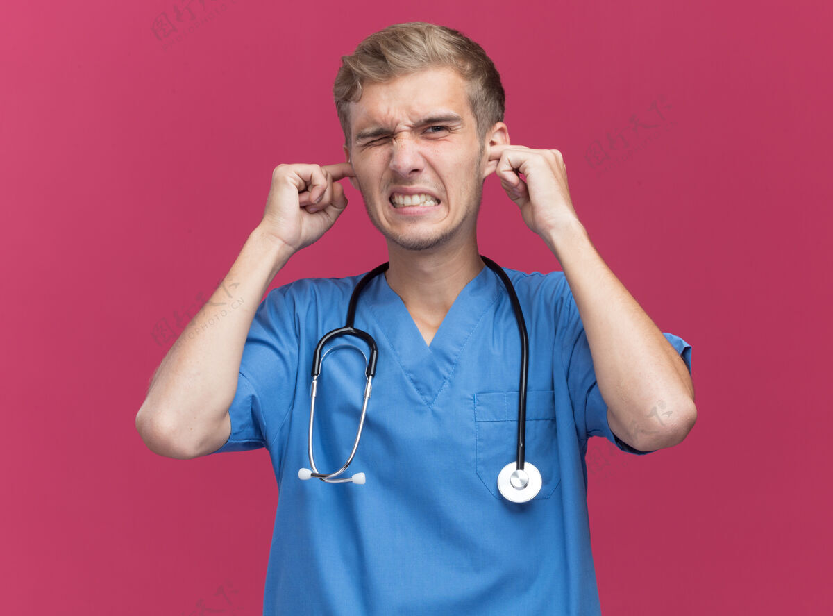 戴恼怒的年轻男医生穿着医生制服 听诊器把耳朵贴在粉红色的墙上听诊器医生男性