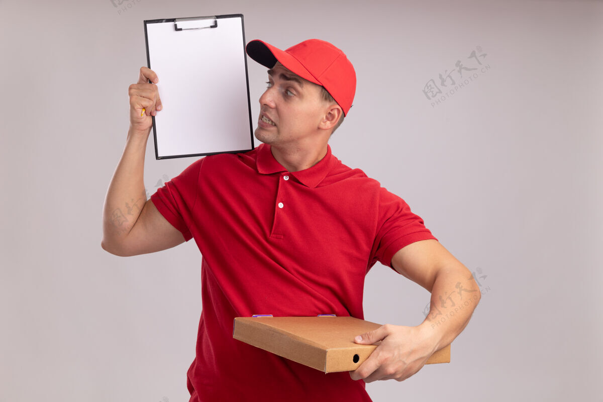 制服关心的年轻送货员穿着制服 戴着帽子 拿着披萨盒 看着他手上的剪贴板孤立在白色的墙上拿着盒子帽子