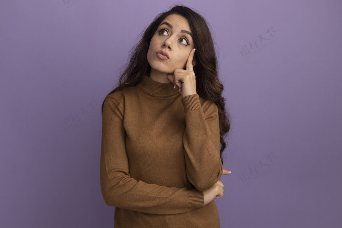 高领毛衣印象深刻的年轻漂亮女孩穿着棕色高领毛衣把手指放在脸颊上隔离在紫色的墙上手指毛衣女孩