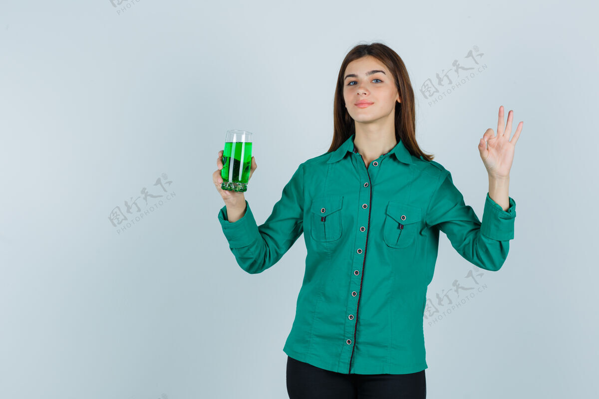 青少年年轻的女孩拿着一杯绿色液体 穿着绿色上衣 黑色裤子 看起来很高兴正面图手持成人秀