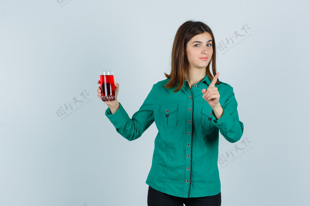 显示年轻女孩拿着一杯红色液体 穿着绿色上衣 黑色裤子 摆出警告的姿态 看起来很可爱 正前方的景色休闲前面玻璃