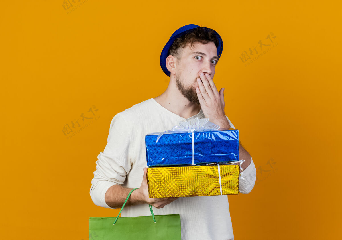 手惊讶年轻英俊的斯拉夫党人戴着党的帽子拿着纸袋和礼品盒手放在嘴上看着相机孤立的橙色背景与复制空间家伙橙色嘴