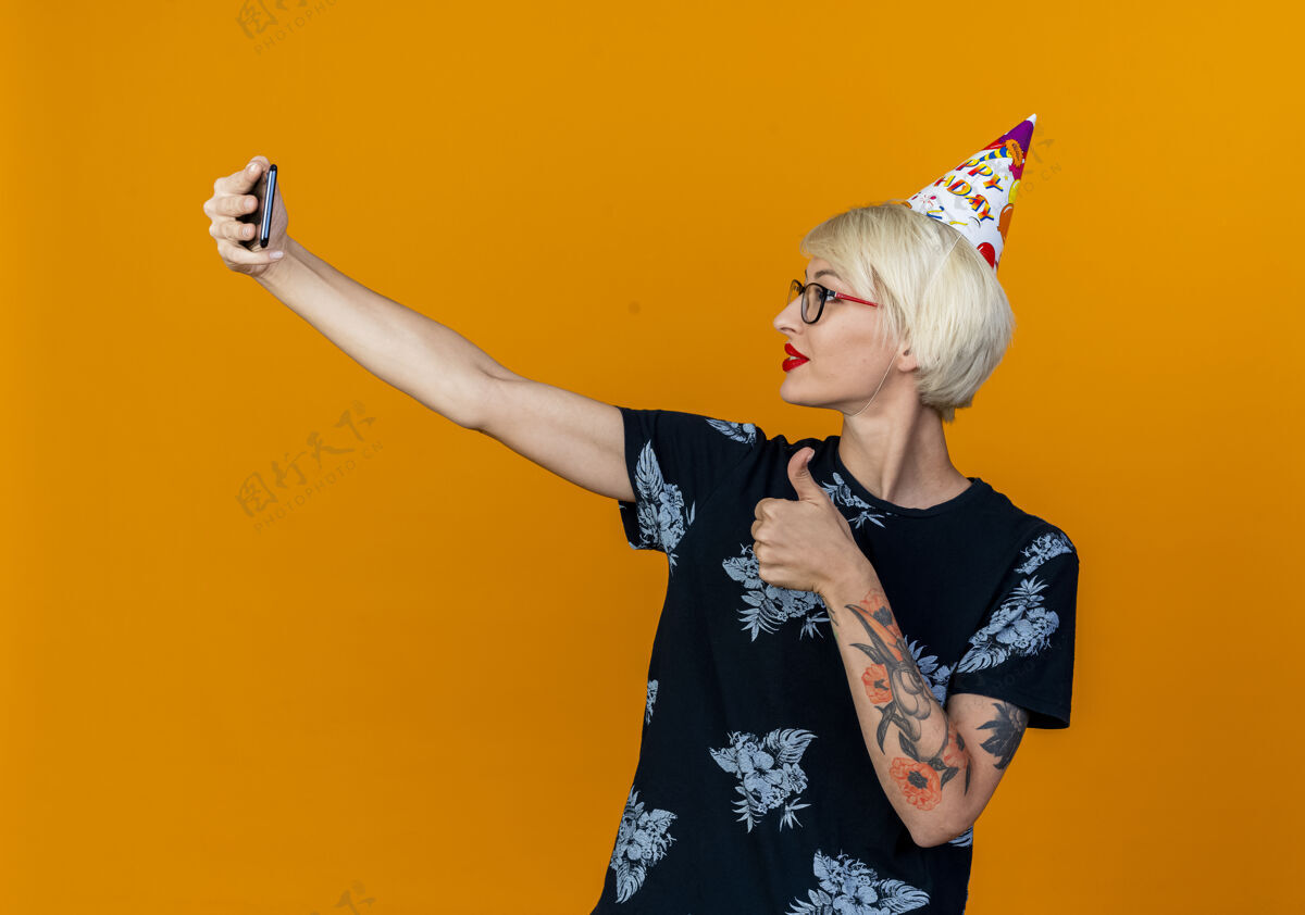 生日年轻的金发派对女孩戴着眼镜和生日帽自拍 在橙色背景下孤立地竖起大拇指女孩请拿
