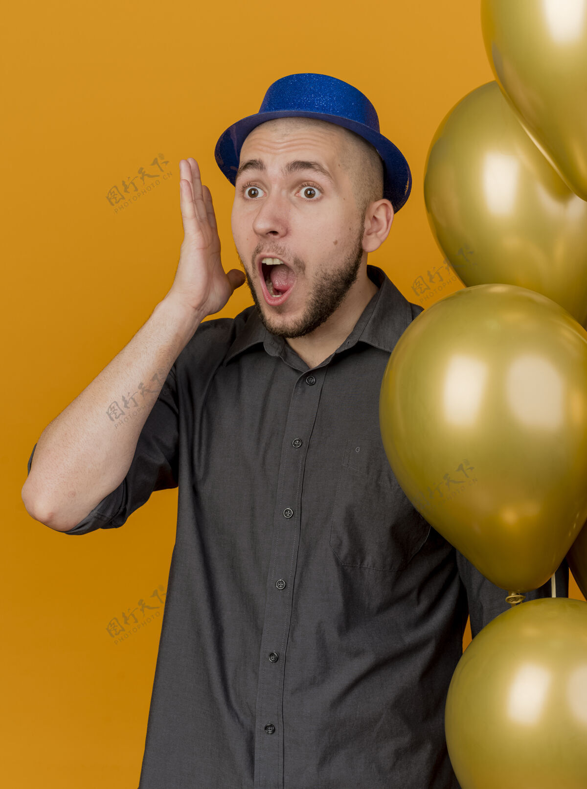直震惊年轻英俊的斯拉夫党的家伙戴着党的帽子拿着气球保持手靠近头部看直橙色背景孤立小伙子手斯拉夫