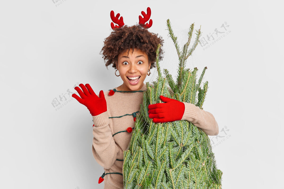 驯鹿节日购物理念积极向上的民族妇女穿着红驯鹿鹿角和手套 挥手致意 在街市上选择圣诞树 在家里装饰新年快乐背心快乐你好