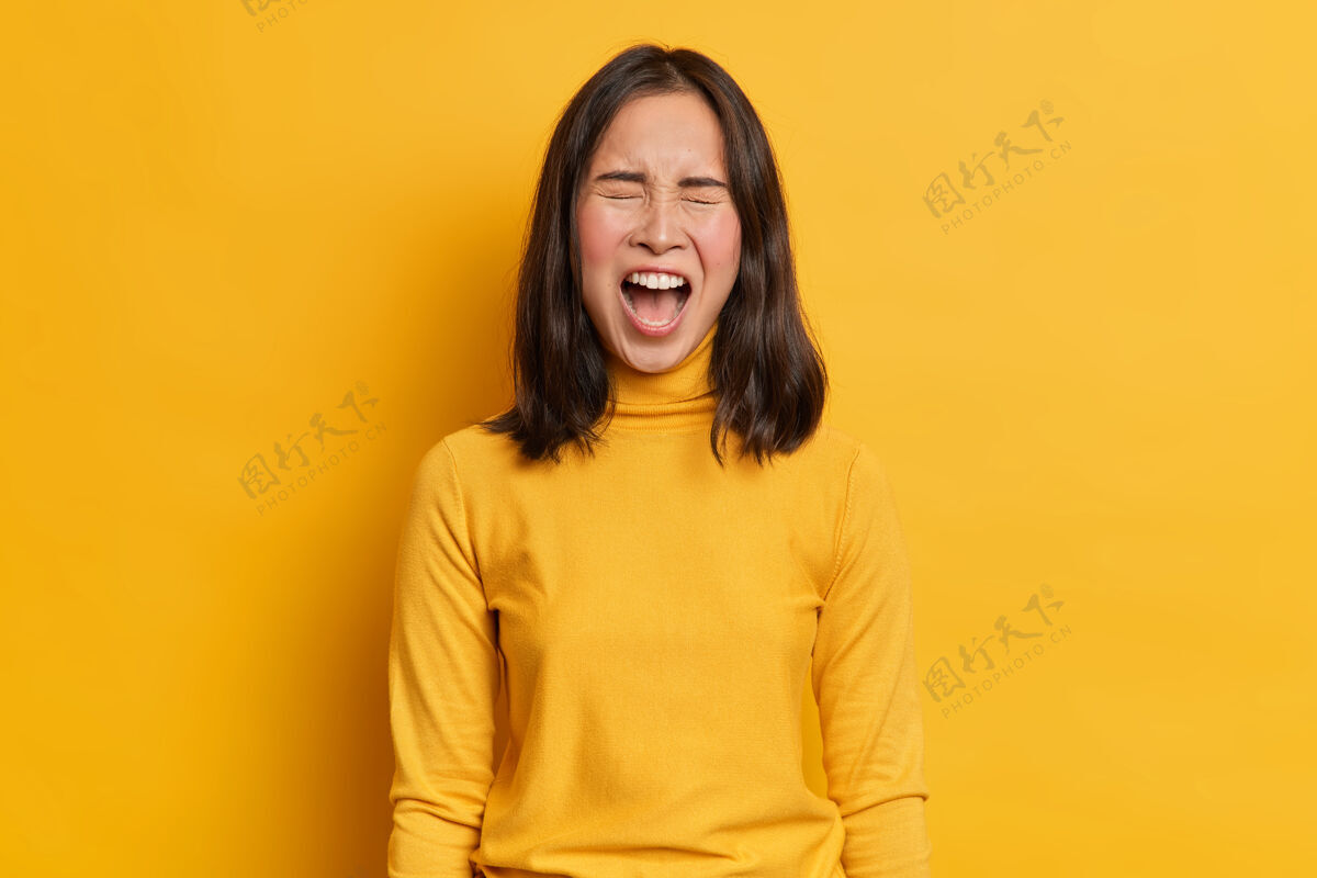 大声情绪化的黑发亚洲女人大声尖叫表达愤怒张大嘴巴穿着休闲的黄色套头衫在同一色调与工作室背景人类的情感和感情的概念压力暴力不满意