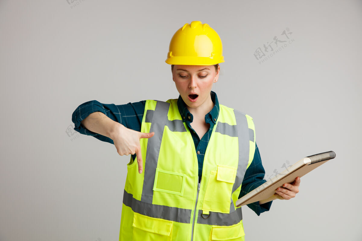 工人惊讶的年轻女建筑工人戴着安全帽和安全背心拿着记事本看着并指着下面指着建筑年轻