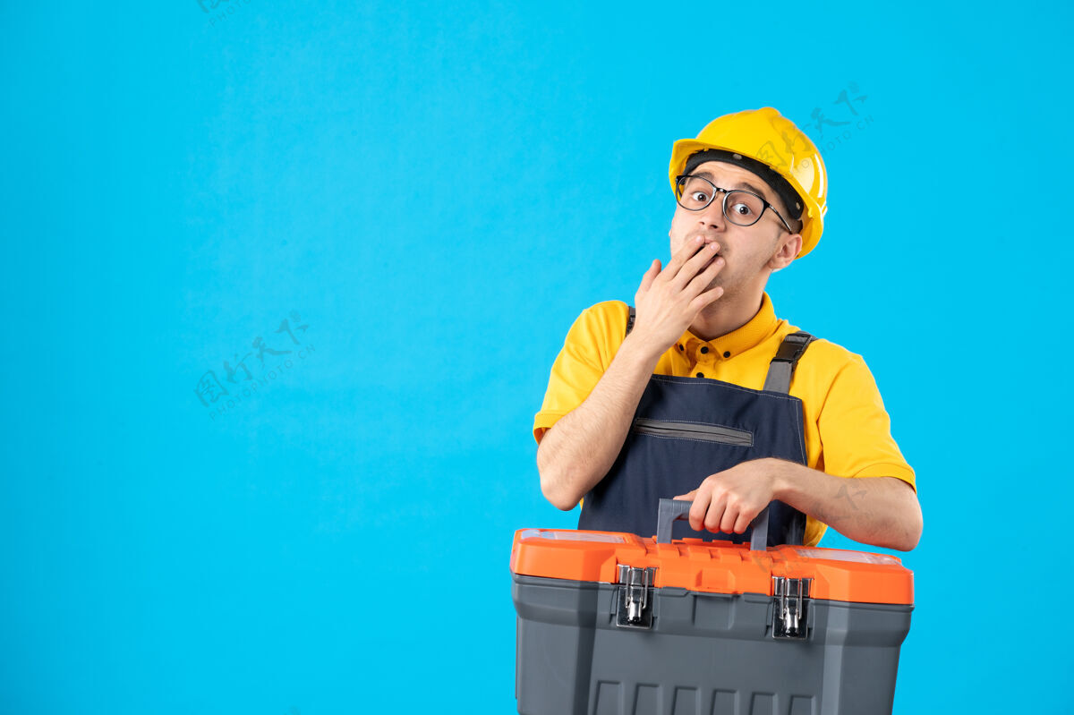 制服身着黄色制服 手持蓝色工具箱的惊讶男工人的正视图箱子建筑工人头盔