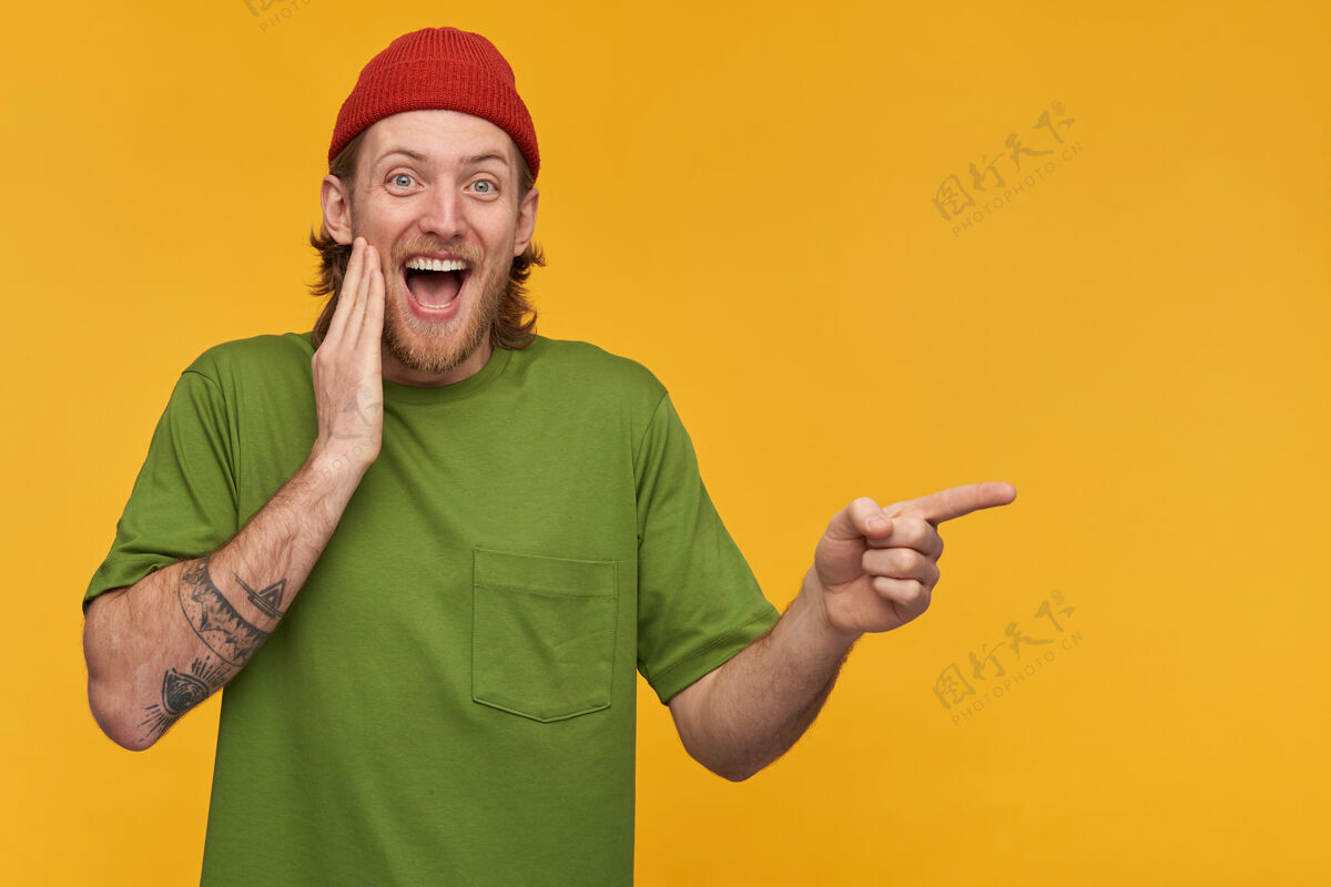胡须笑容可掬的金发胡子 穿着绿色t恤和红色小珠子 身上有纹身手指指向右边的复制空间 隔离在黄色的墙上年轻触觉情绪