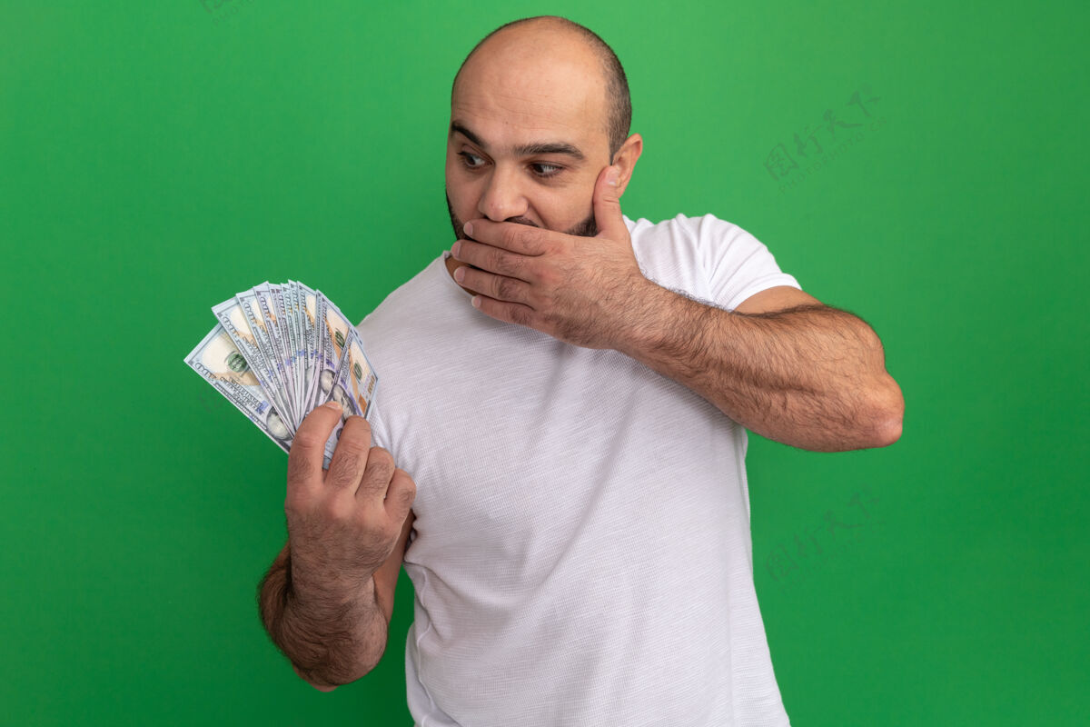胡子一个留着胡须 穿着白色t恤的男人拿着现金看着钱 惊讶地捂着嘴 手站在绿色的墙上掩护惊喜惊喜