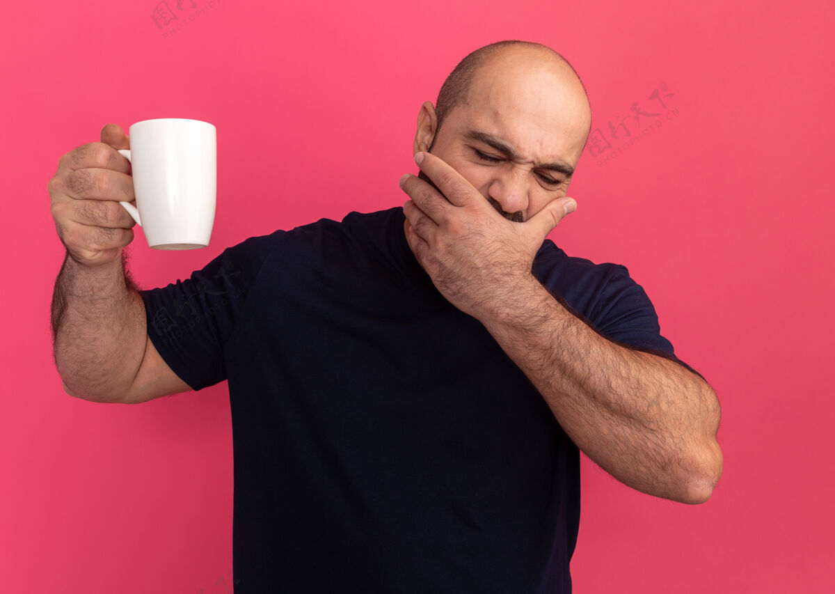 掩护一个留着胡子的男人 穿着海军蓝t恤 手里拿着一个杯子 看起来很累 手放在粉红色的墙上 捂住嘴杯子站着累了