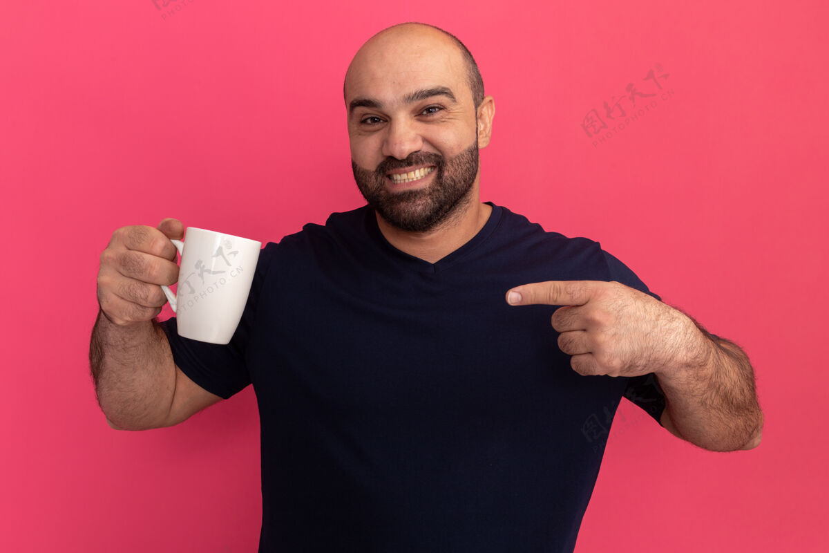 站身穿海军蓝t恤的大胡子男人拿着一个杯子 用食指指着杯子 站在粉红色的墙上 开心而积极地微笑着手指正面胡子