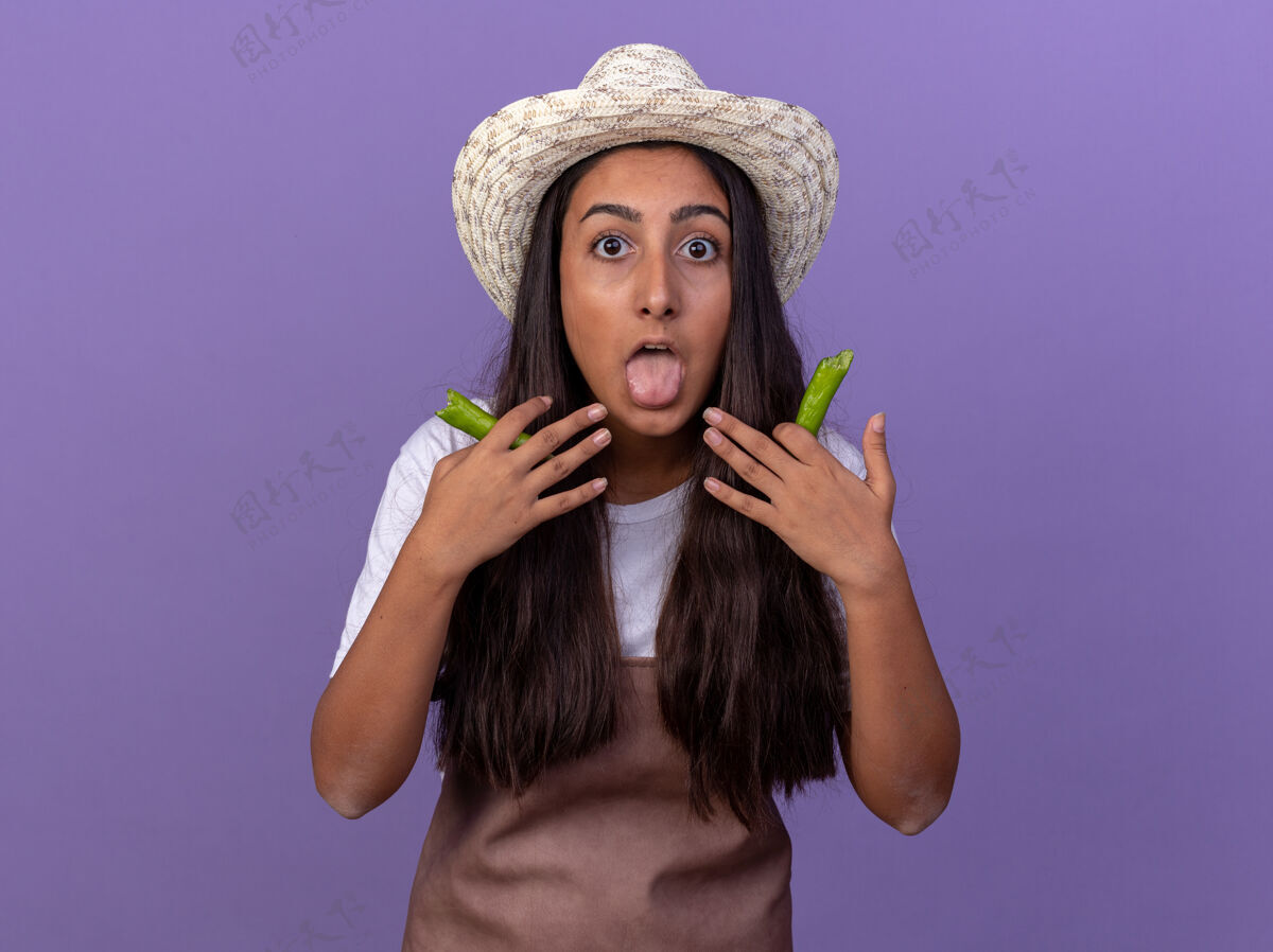辣椒穿着围裙 戴着夏帽 手里拿着青椒的年轻园丁女孩 心急如焚地伸出舌头站在紫色的墙上辣椒年轻站