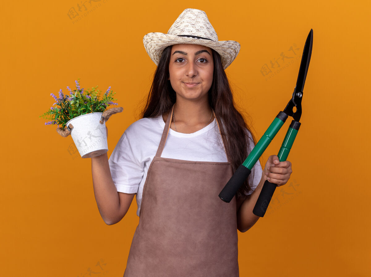剪子穿着围裙 戴着夏帽 手持树篱剪和盆栽的年轻园丁女孩站在橙色的墙上 脸上带着微笑年轻围裙微笑