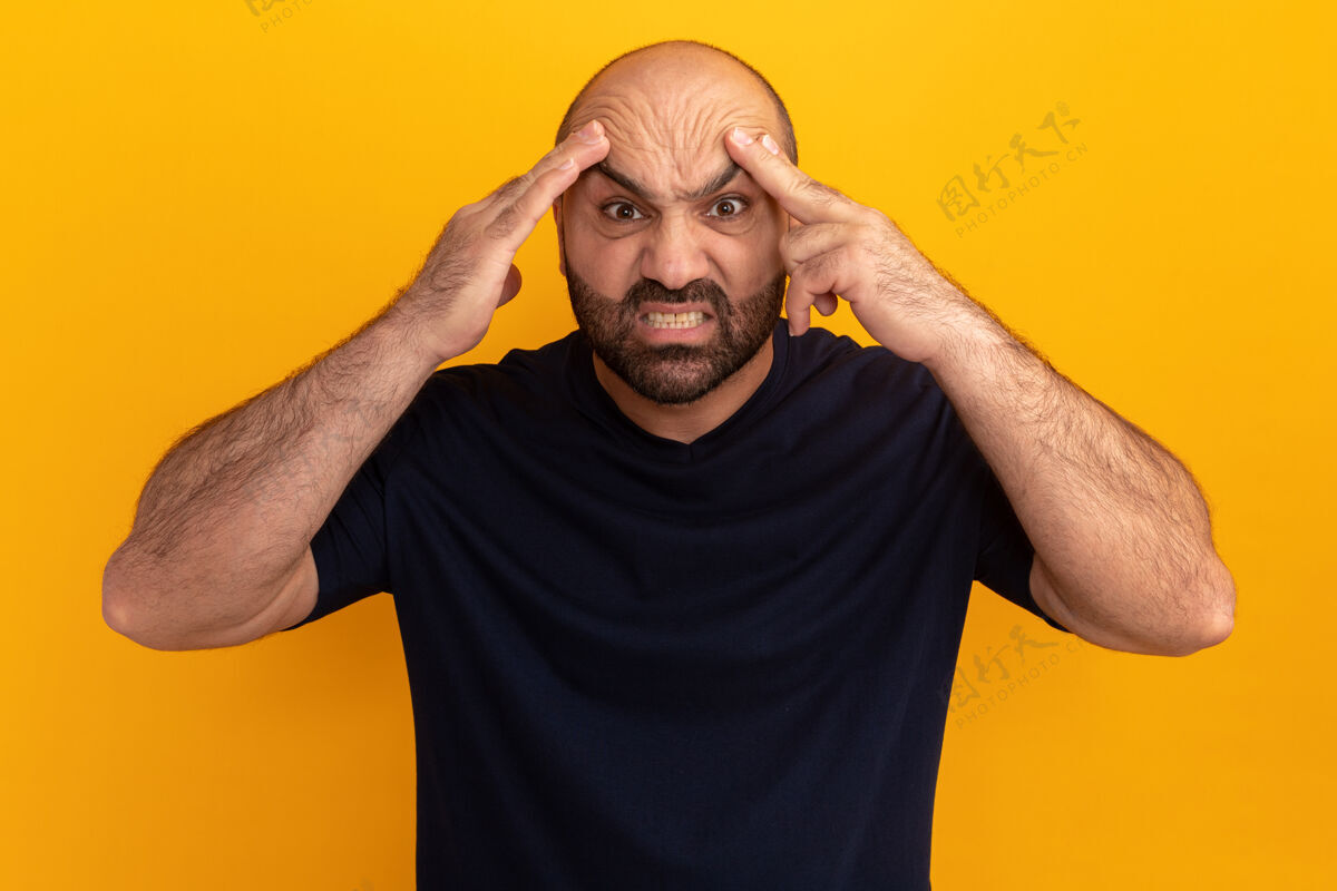 胡子一个留着胡子的男人 穿着海军蓝t恤 愤怒的脸碰到站在橙色墙上的头站生气男人