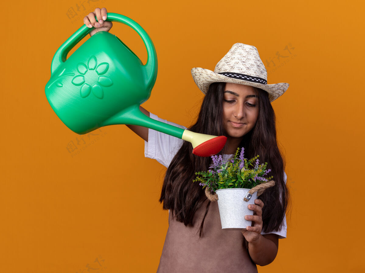微笑快乐的年轻园丁女孩 围裙戴着夏帽 手里拿着浇灌罐 脸上带着微笑的盆栽浇灌植物站在橙色的墙上锅围裙帽子