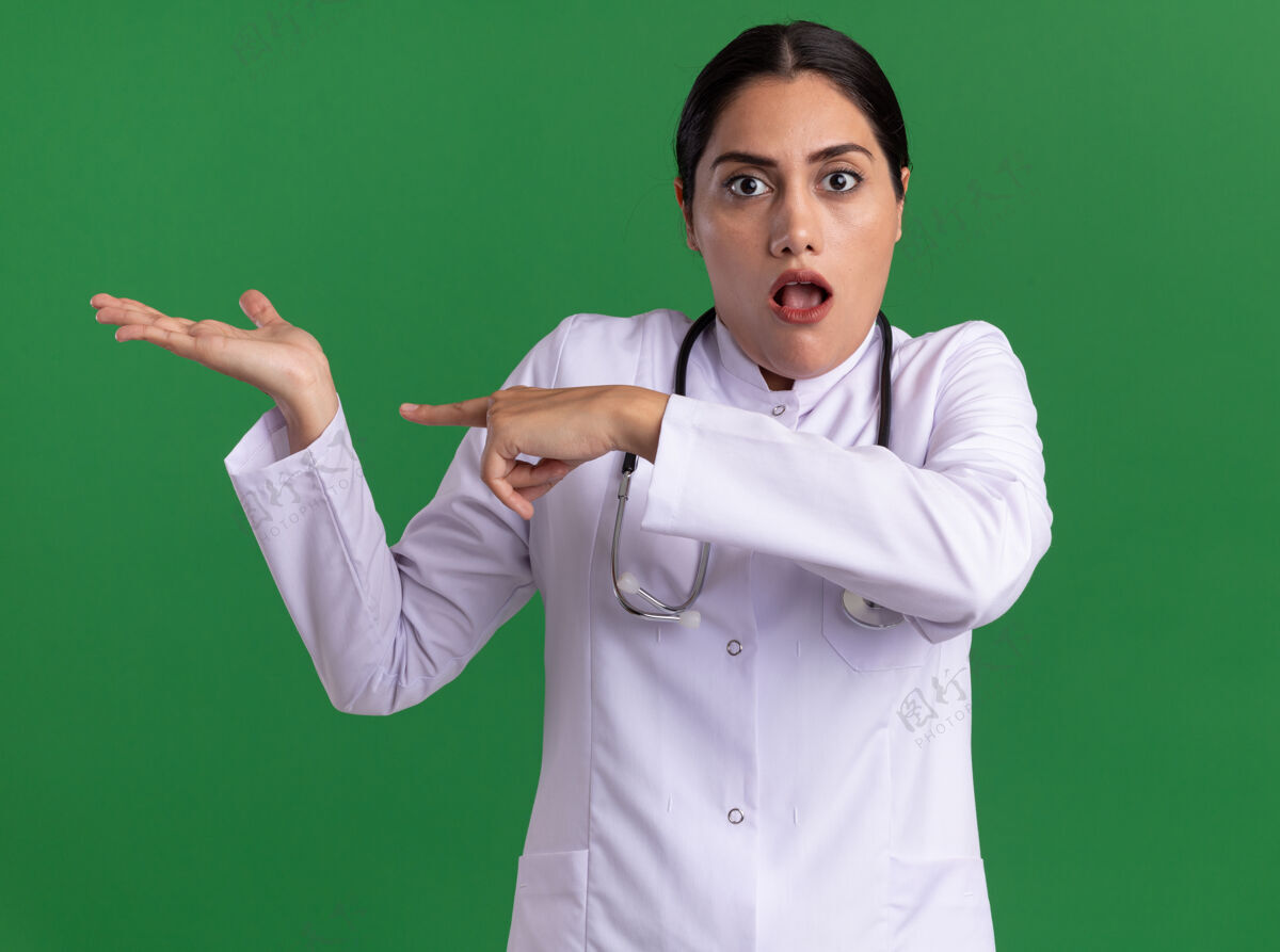 医生年轻的女医生穿着医用外套 脖子上戴着听诊器 她惊讶地用食指指着绿墙上的一边指着惊喜周围