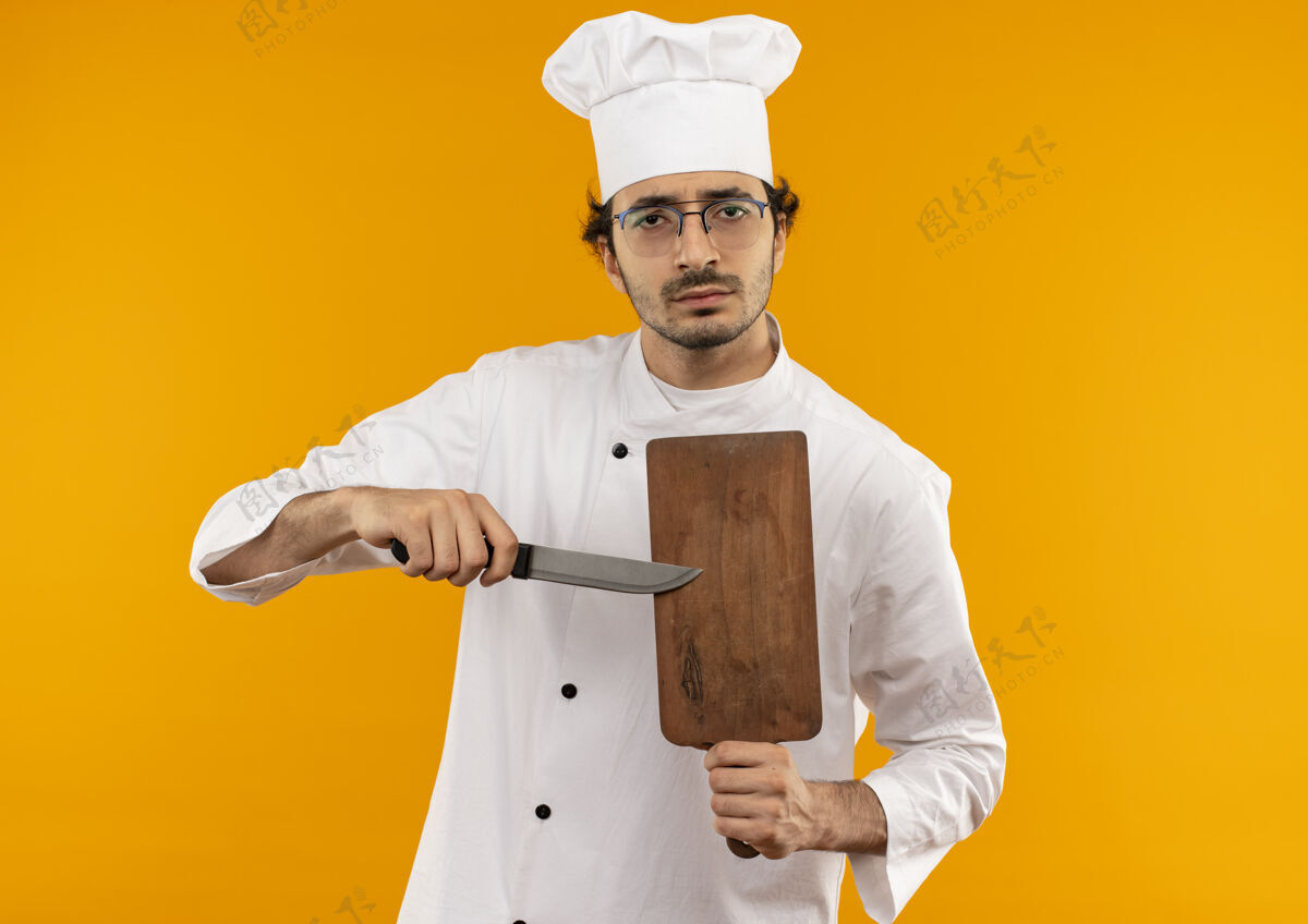 板自信的年轻男厨师穿着厨师制服 戴着眼镜 手里拿着砧板和刀切割制服刀