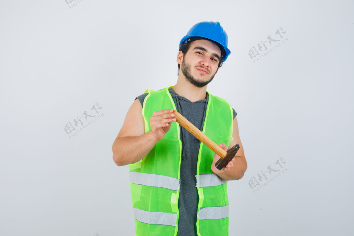 年轻年轻的建筑工人穿着工装展示锤子 看起来很高兴 正面视图微笑帽子男性