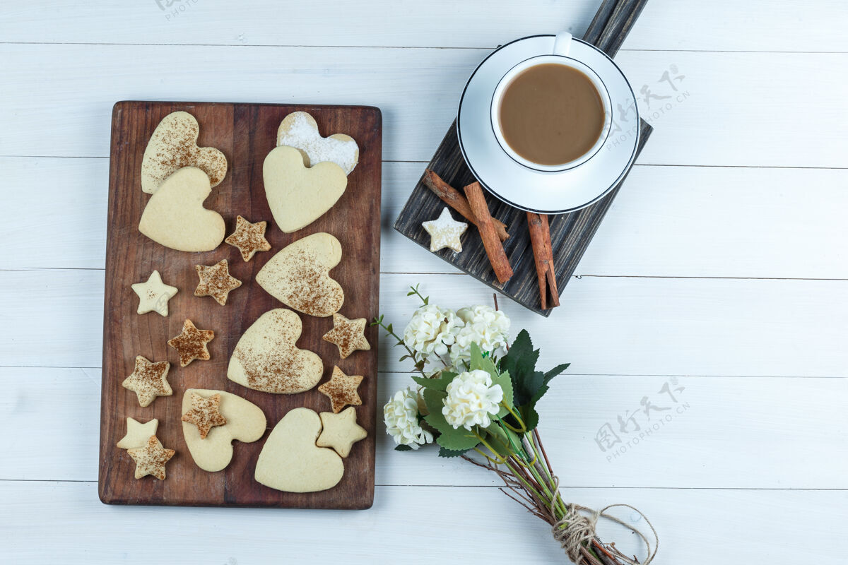 饼干心形和星型饼干放在一个木制的砧板上 上面放着一杯咖啡 鲜花 肉桂平放在一块白色的木板背景上婚礼奶油烘焙