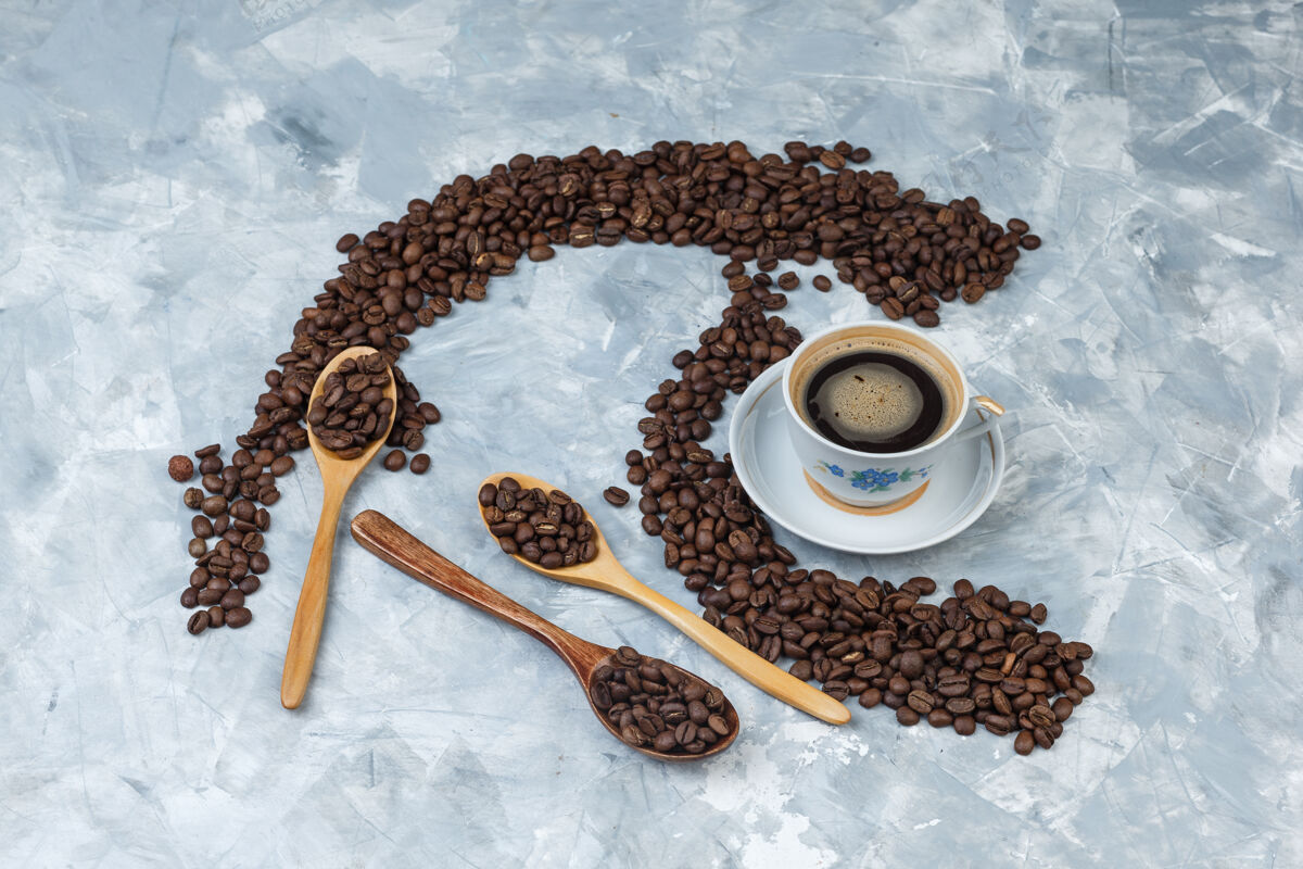 杯子一些咖啡豆和咖啡饮料放在杯子里 木制勺子放在灰色的石膏背景上 平放自然泡沫种子