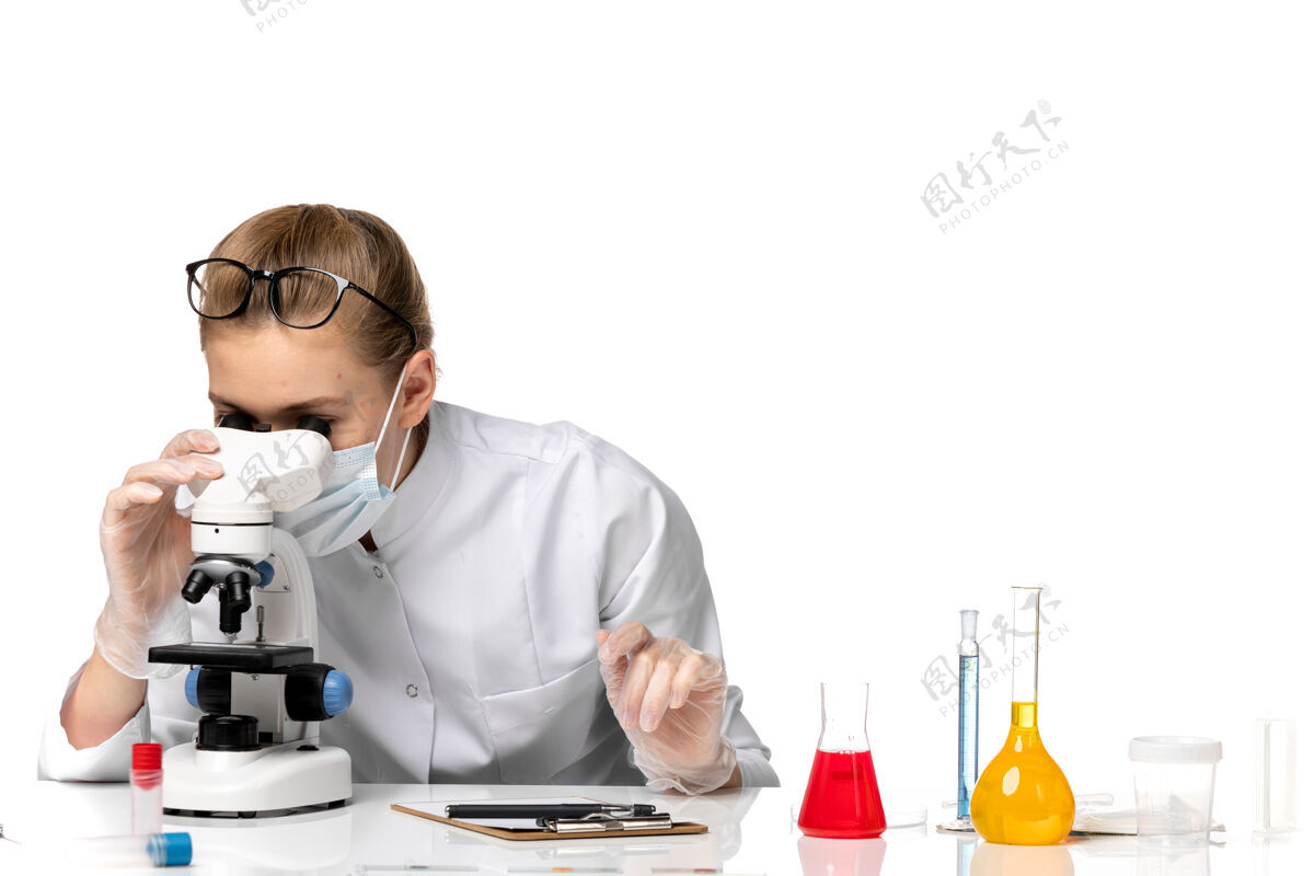 显微镜前视图女医生穿着医疗服戴着口罩 由于戴着隐形眼镜 用显微镜在浅白的空间里观察病毒到期大流行