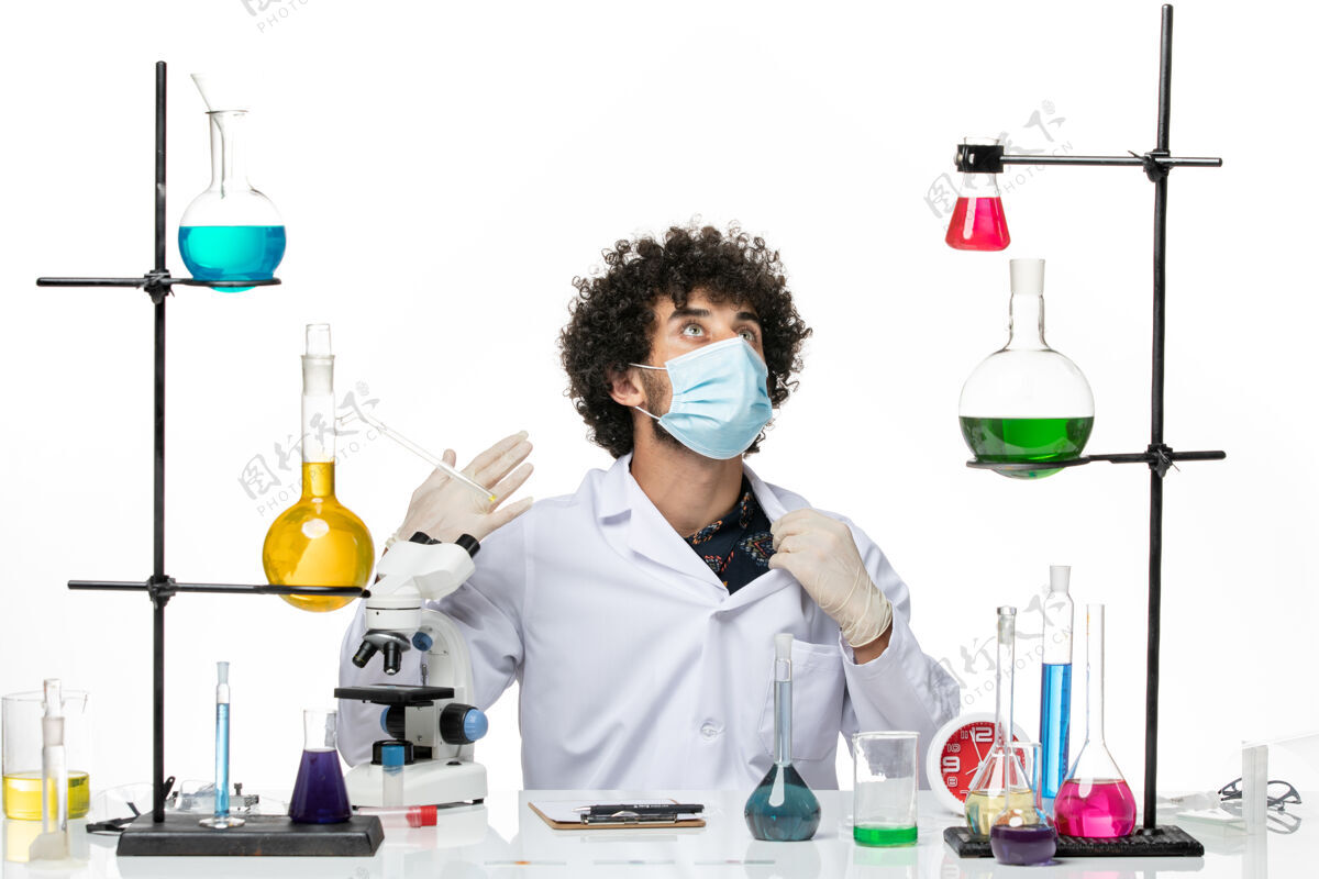 面具前视图男性化学家穿着医疗服 戴着面罩 坐在浅白的空间里 拿着溶液男性化学家实验室烧杯