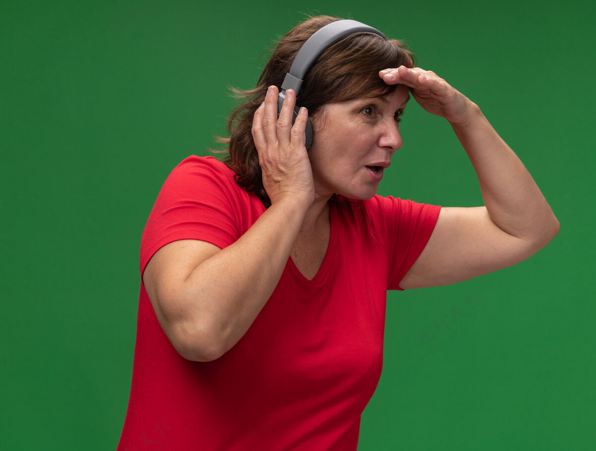 远穿着红色t恤的中年妇女戴着耳机望着远处 手举着头站在绿色的墙上女人中头