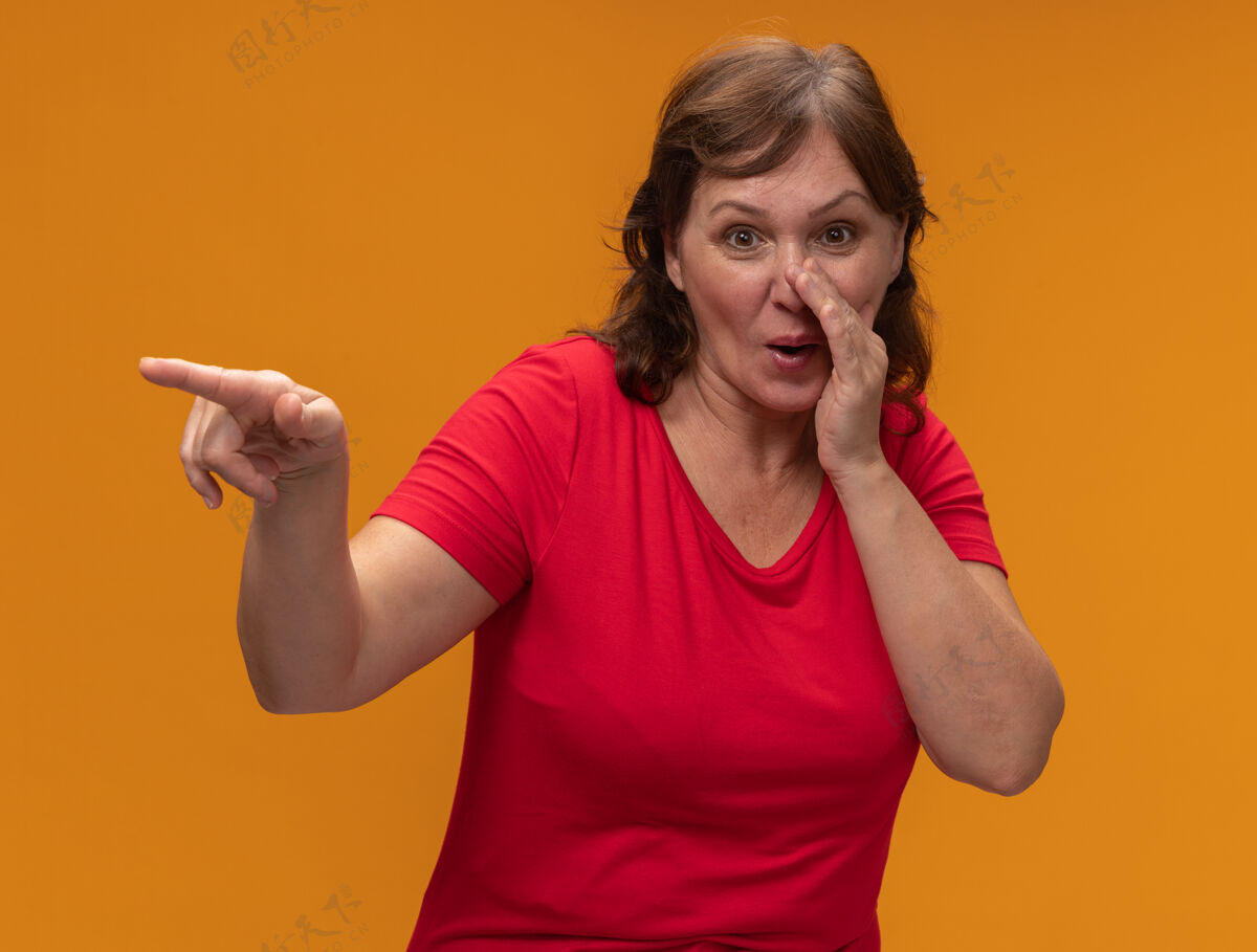 附近身穿红色t恤的中年妇女微笑着说秘密 手靠近嘴巴 食指指向站在橙色墙上的东西站什么手指