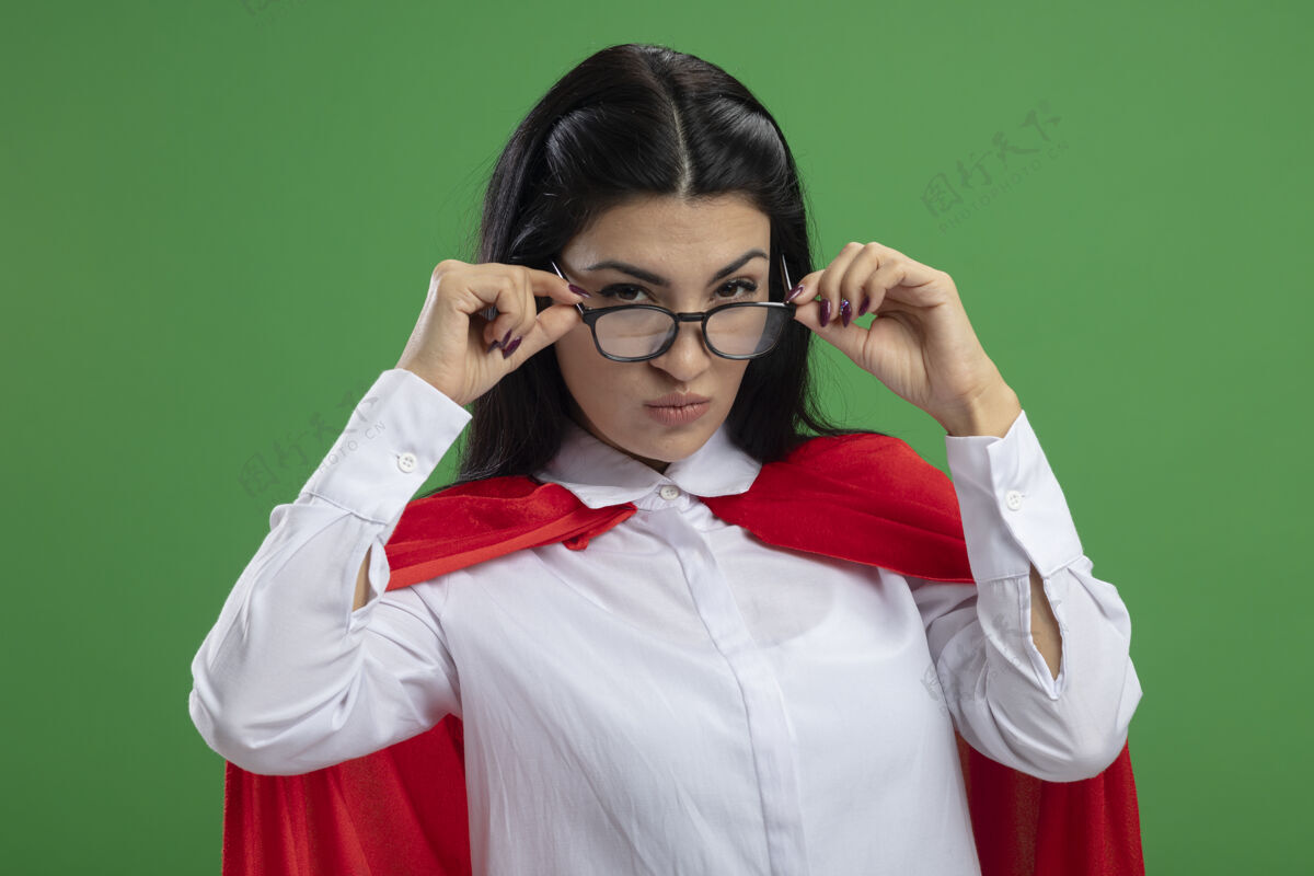 狡猾严肃狡猾的年轻白种人超级英雄女孩戴着眼镜手拿眼镜可疑的神情孤立在绿色的墙上手穿着女孩