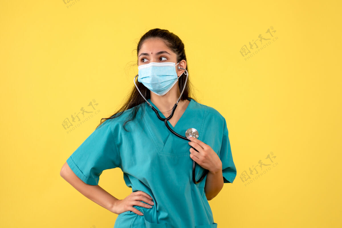 人黄色墙上手持听诊器的戴着医用面罩的年轻女医生的正视图电话医疗护士