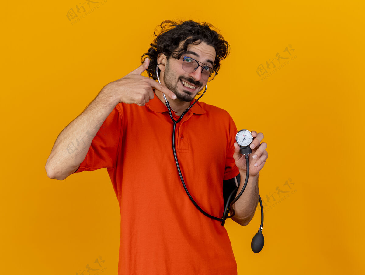 听诊器微笑着的年轻白种人 戴着眼镜和听诊器 拿着血压计 指着隔离在橙色墙壁上的血压计 并留有复印空间橙色指尖男人