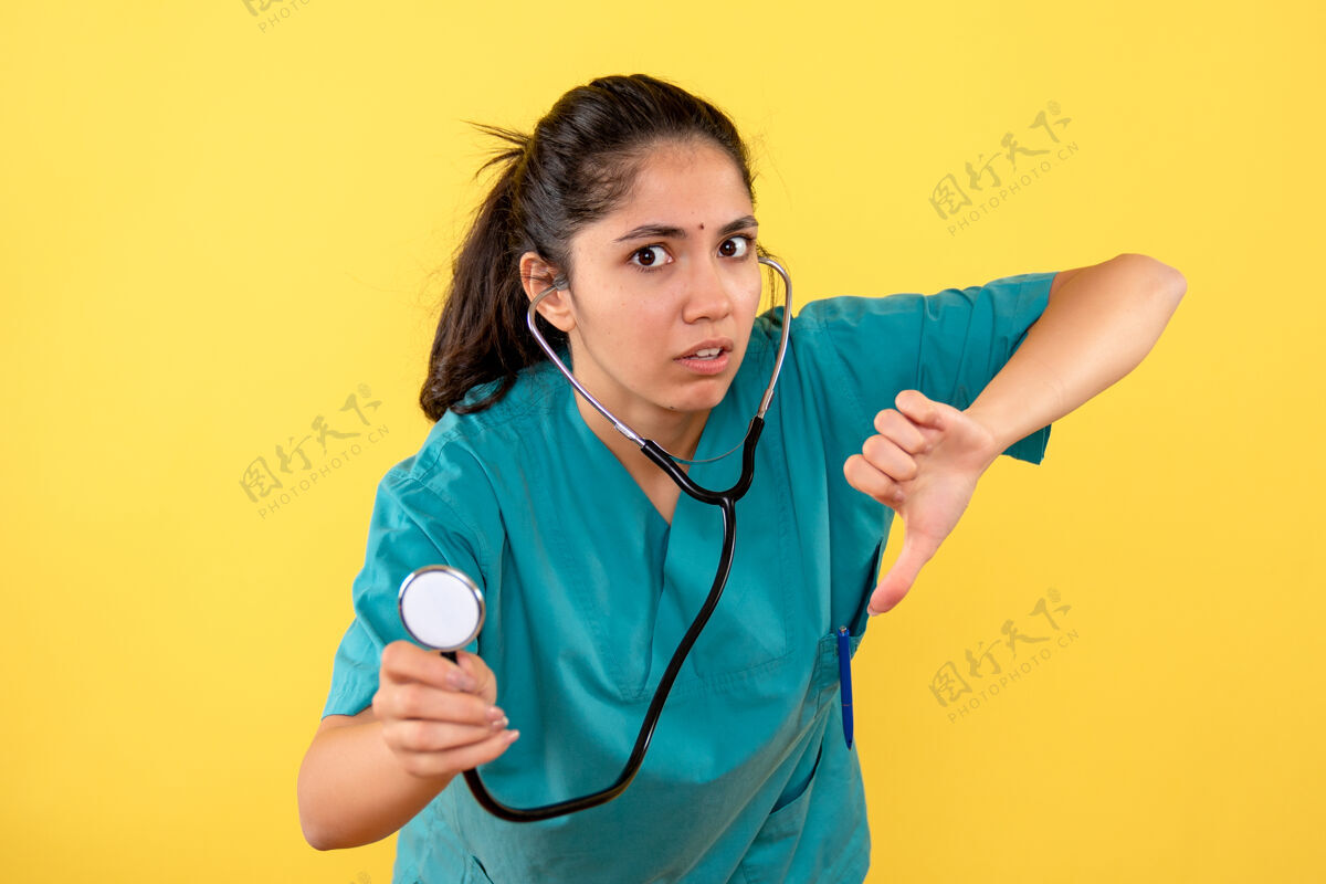 前面身穿制服的女医生正拿着听诊器在黄色墙上做拇指朝下的手势医疗护理护士