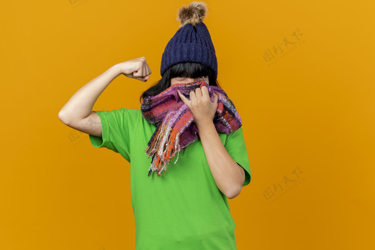 年轻自信的年轻病高加索女孩戴着冬天的帽子和围巾 用围巾遮住嘴巴做着强烈的手势 隔离在橙色的墙上 留着复制空间手势橙色帽子