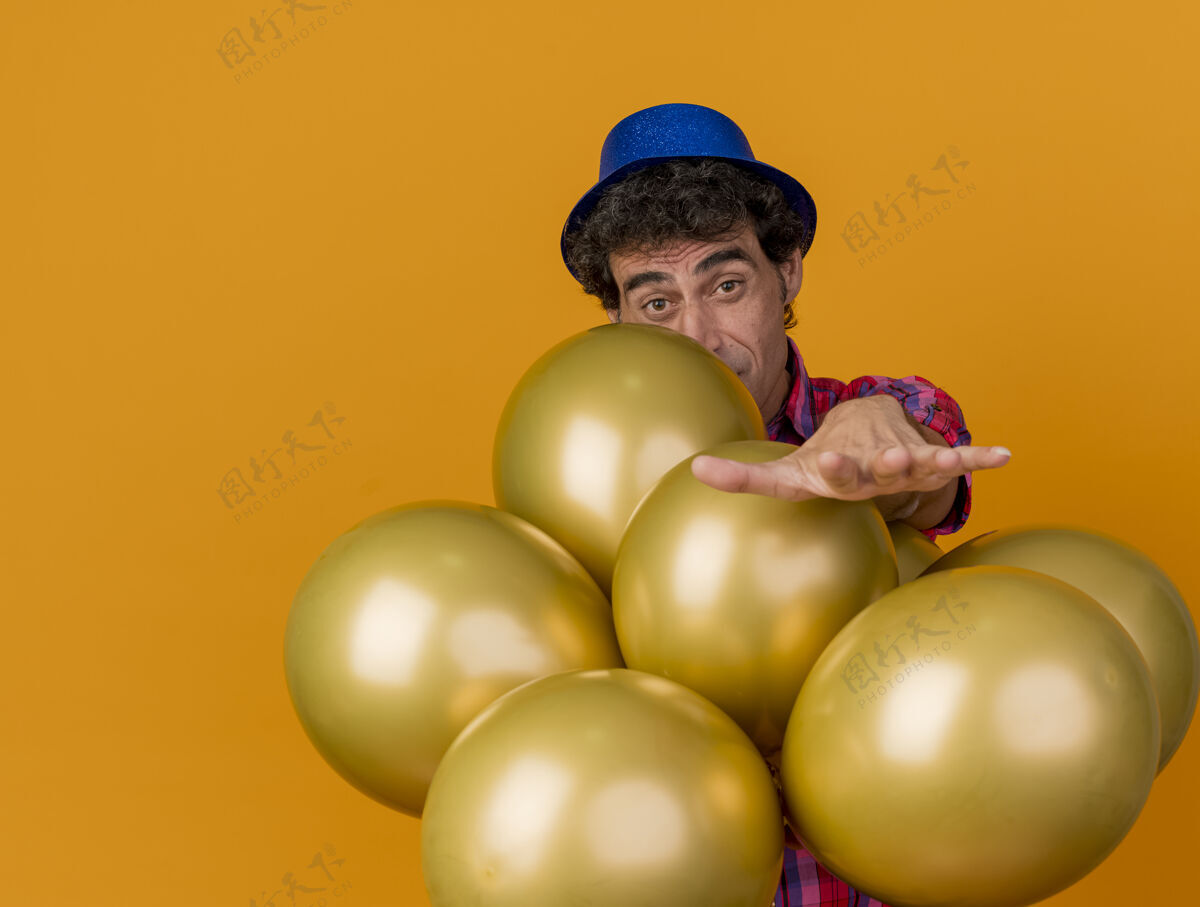帽子印象深刻的中年白种人党男子戴着党的帽子站在气球后面伸出手来孤立在橙色墙上的复制空间中年气球橙色
